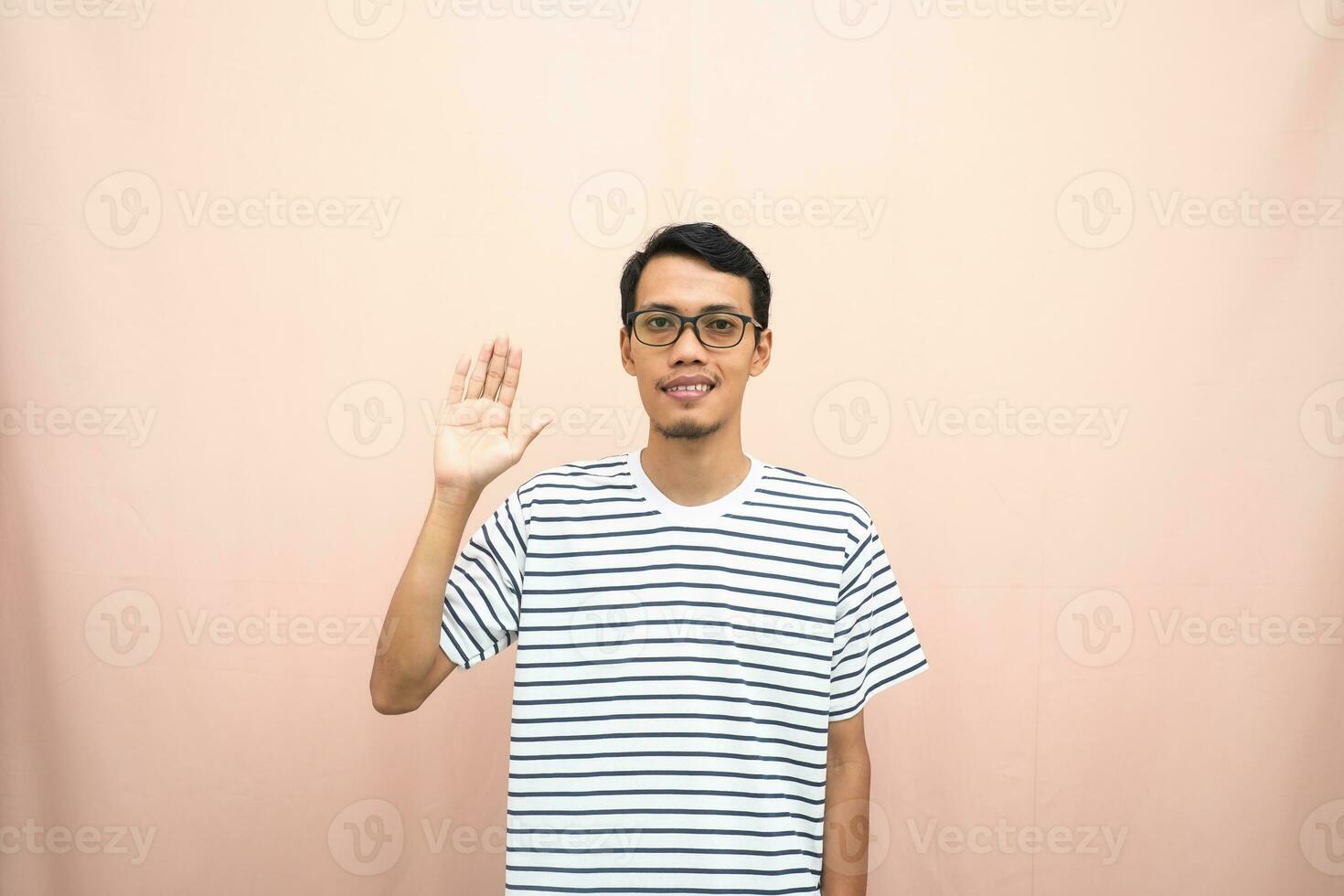 asiático hombre en lentes vistiendo casual a rayas camiseta, saludo actitud y sonriente. aislado beige antecedentes. foto