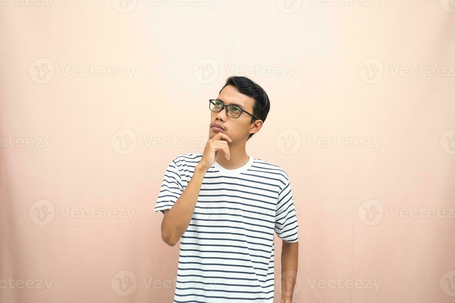 Asian man in glasses wearing casual striped t-shirt, gesturing get ideas, looking for ideas, thinking. Isolated beige background. photo