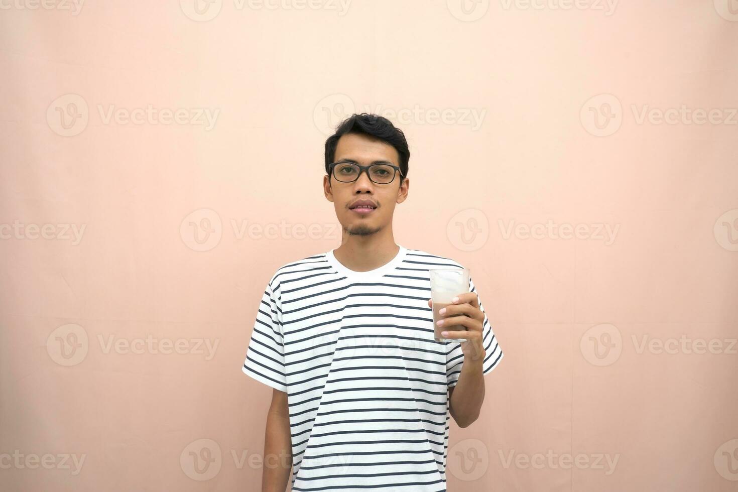 portrait of an asian man wearing glasses wearing a casual striped t-shirt. Enjoying drinking chocolate milk. Isolated beige background. photo