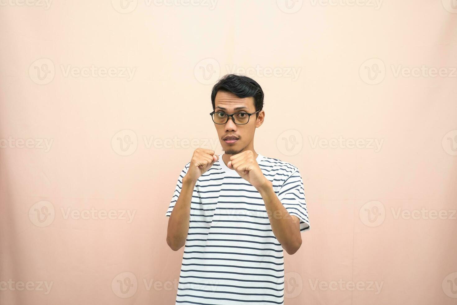 Asian man with glasses wearing casual striped shirt, fist fist pose, indicating strong or ready to fight. Isolated beige background. photo