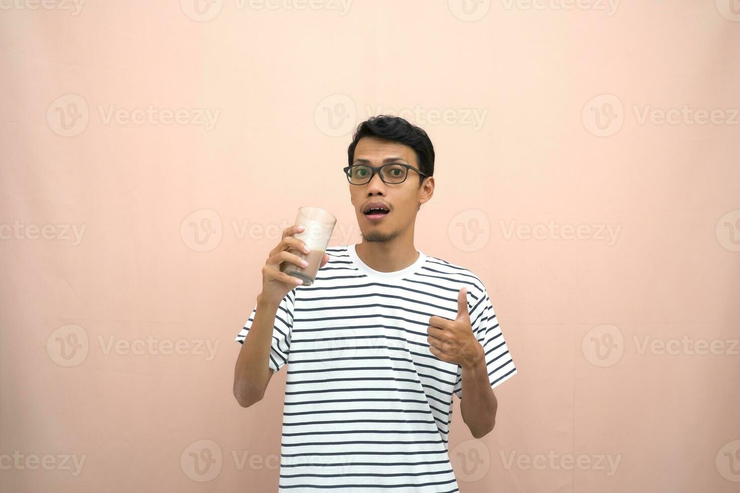 portrait of an asian man wearing glasses wearing a casual striped t-shirt. Enjoying drinking chocolate milk. Isolated beige background. photo