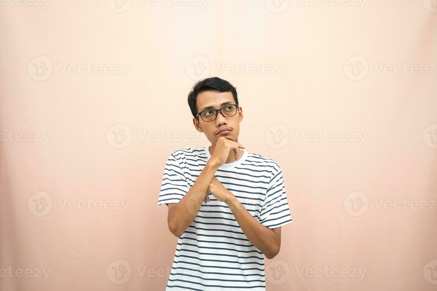 Asian man in glasses wearing casual striped t-shirt, gesturing get ideas, looking for ideas, thinking. Isolated beige background. photo