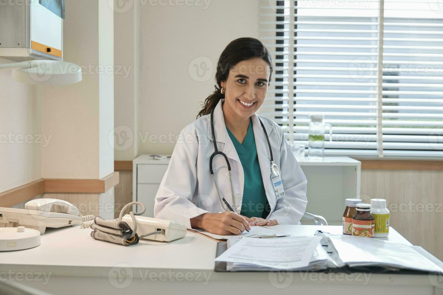 retrato de hermosa hembra médico y profesional nutricionista en uniforme sonriente y mirando a cámara con suplementario comidas en escritorio para sano dieta a hospital. foto