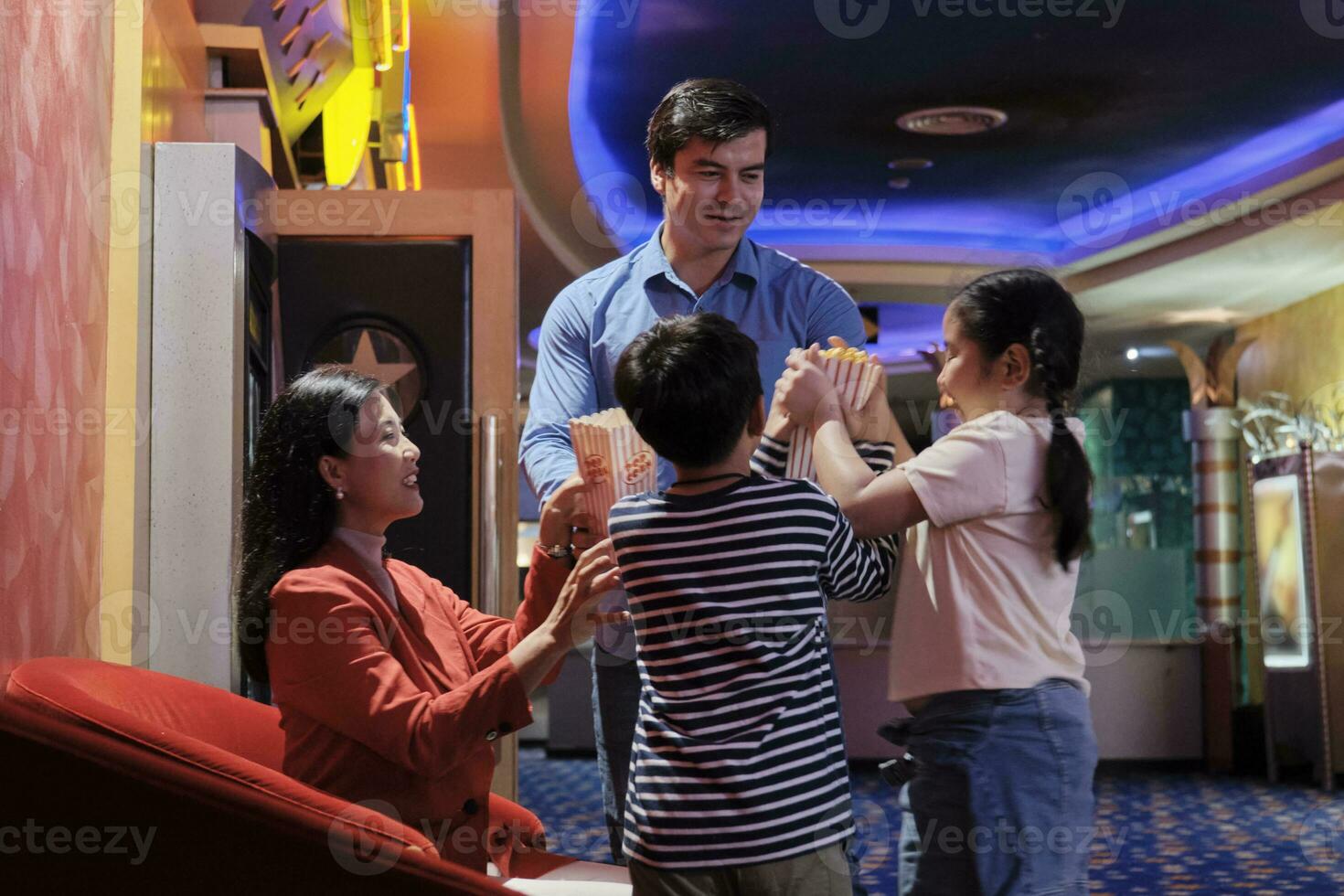 Cheerful Asian family at cinema show. Mum and kids sit in theater seat, waiting for father's popcorn before watching movie, and happy together, a public indoor entertainment lifestyle with film shows. photo
