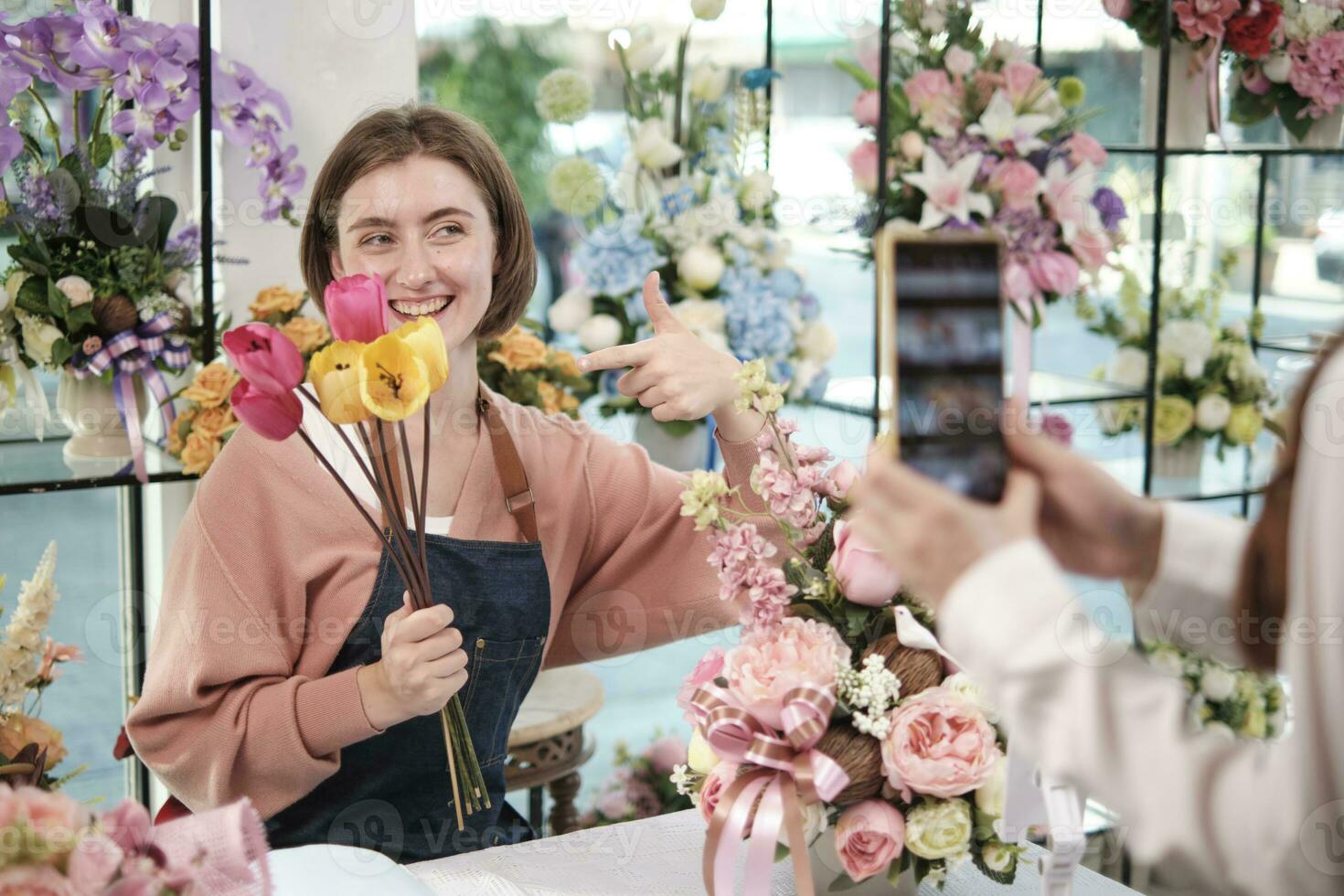 comercio electrónico negocio, uno joven blanco hembra florista demuestra y muestra floral preparativos vía en línea En Vivo transmisión con teléfono inteligente solicitud en brillante flor comercio, hermosa flores almacenar. foto