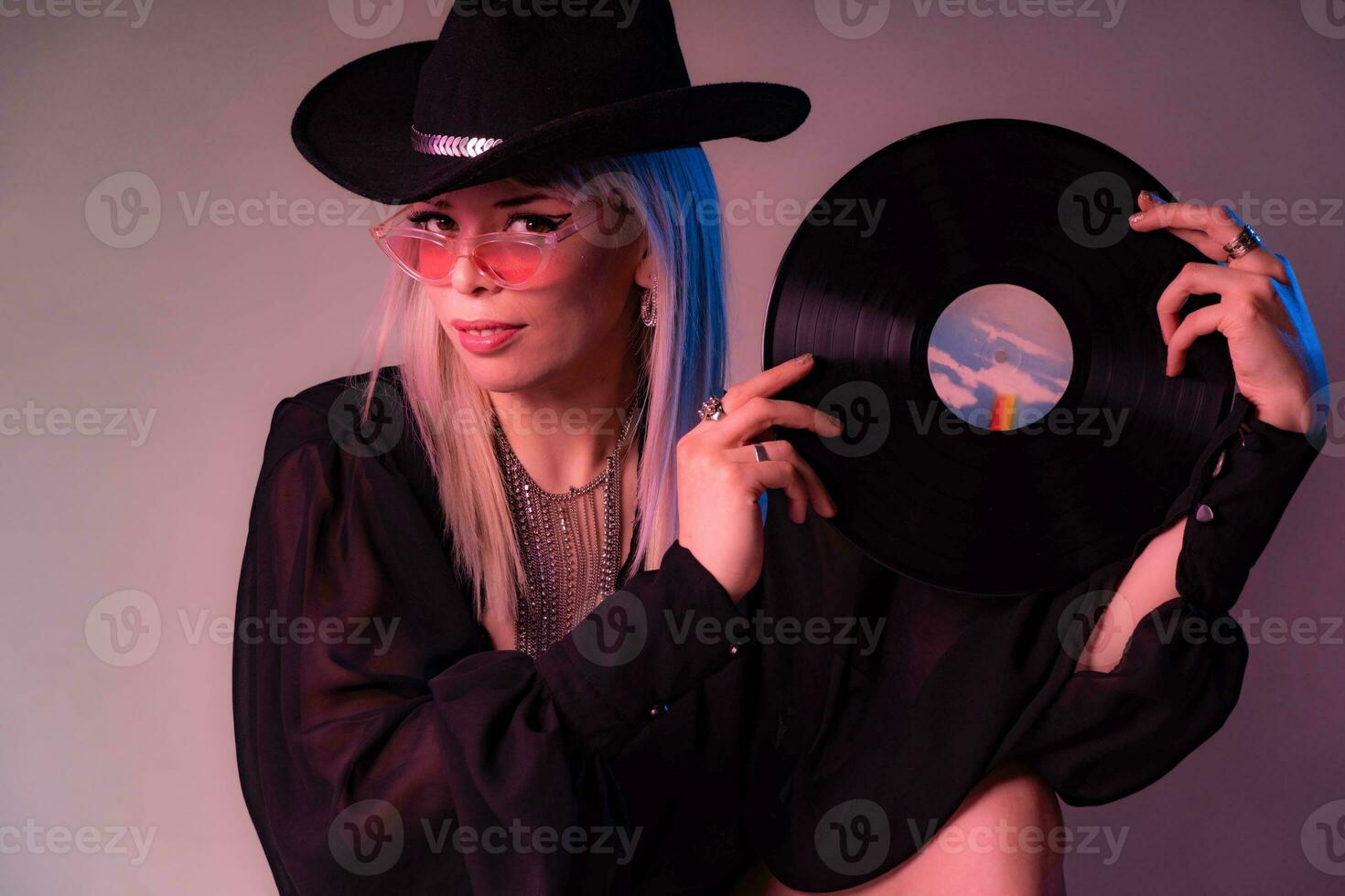 Sexy Dj woman looks over colored glasses holding a vinyl record with a black hat. photo