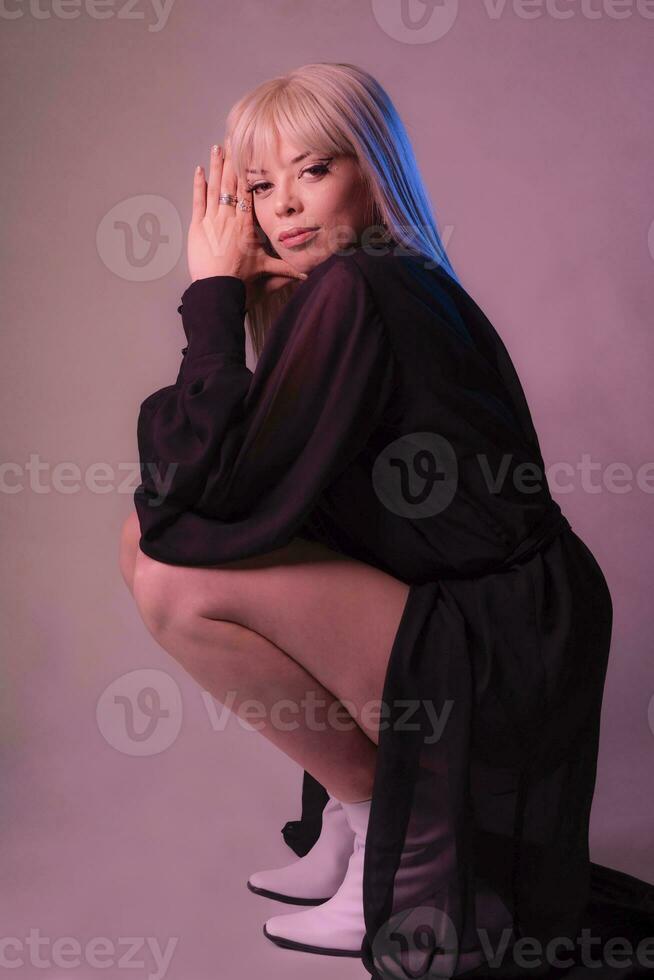 Blond Woman squatting down, wearing a long black dress and white boots. photo