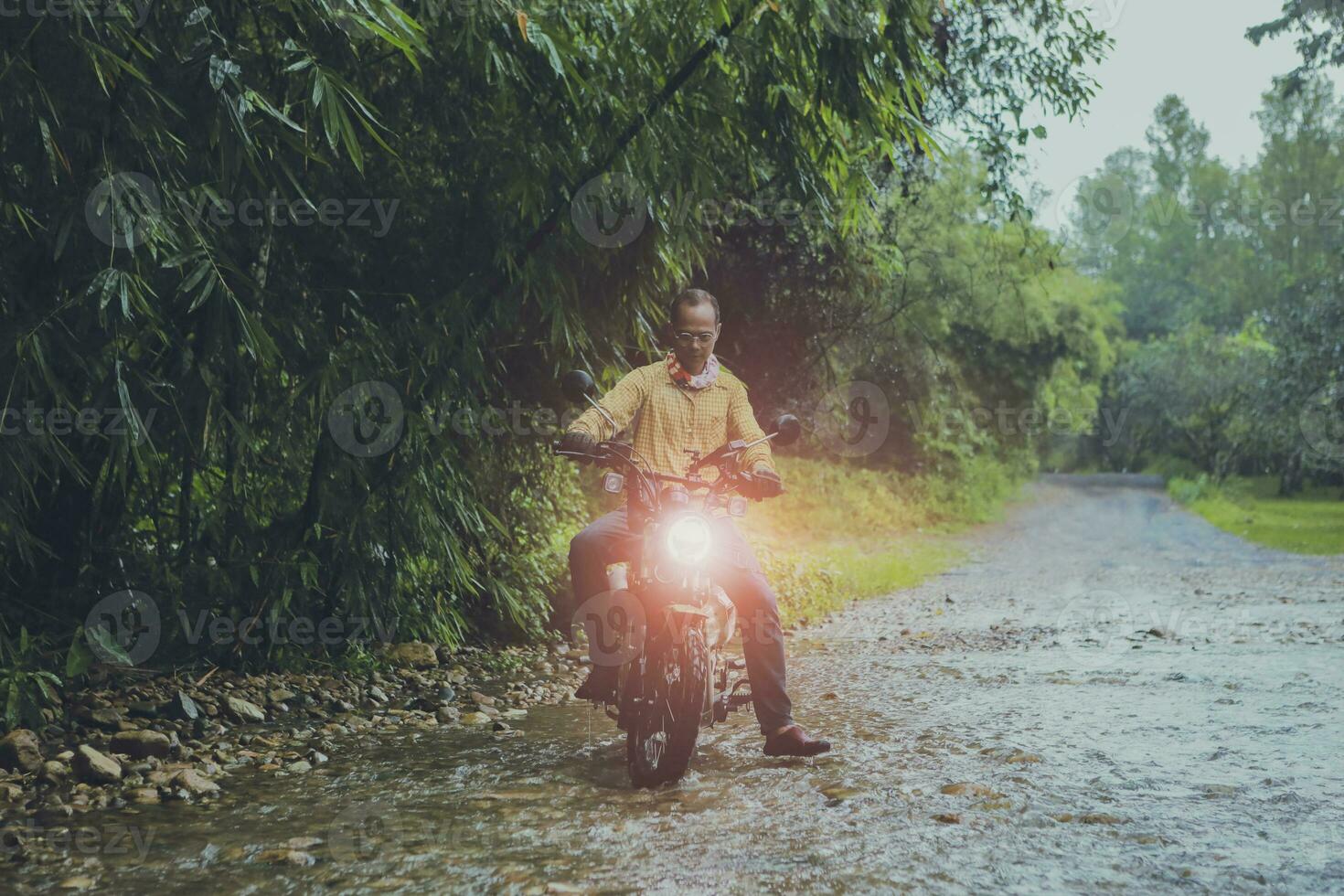 asian man riding small enduro motorcycle crossing flowing creek in forest among rain falling photo