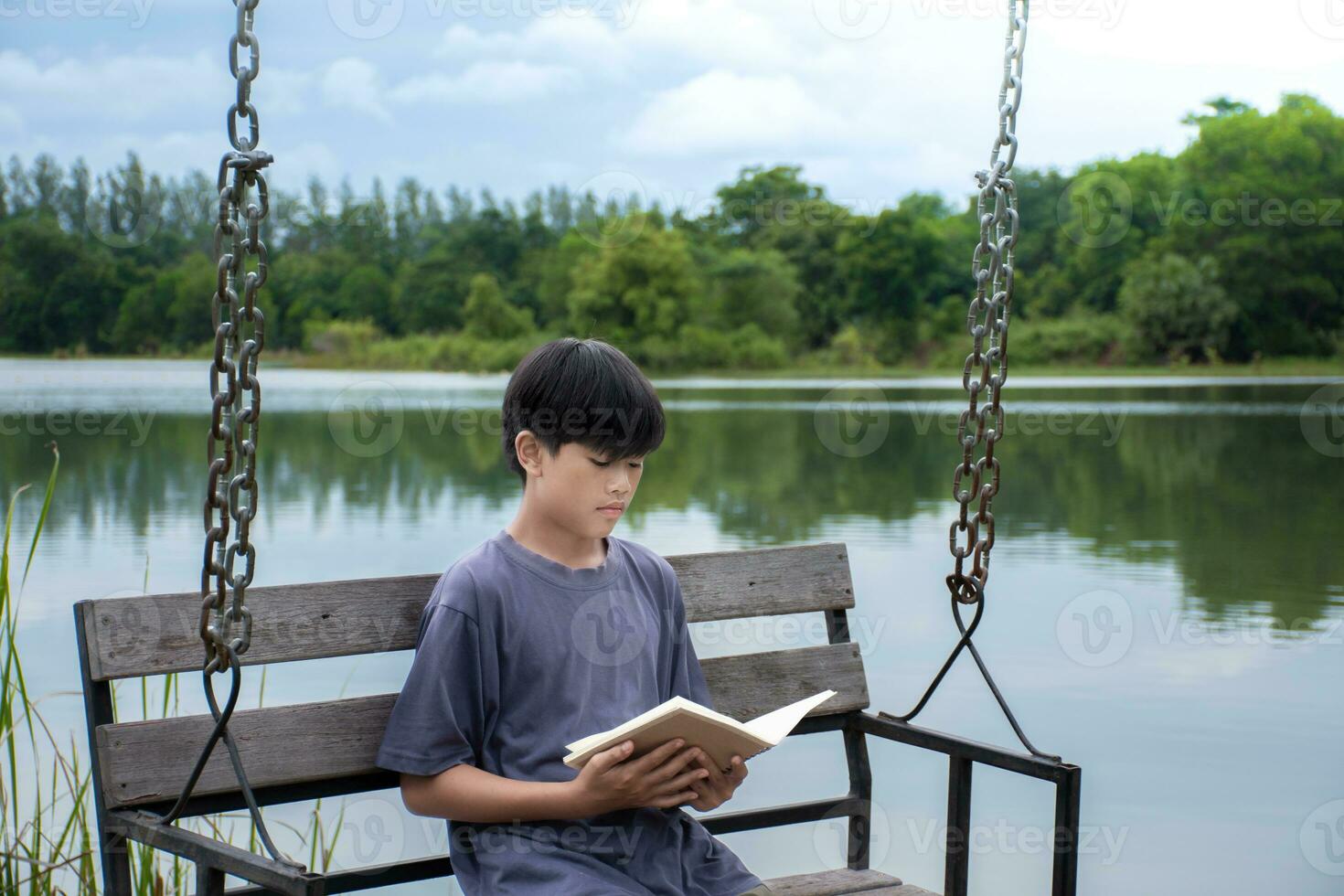 asiático chico sentado leyendo libro en columpio a orilla antecedentes es bosque y cielo con amanecer, chico azul camisa, niños con libro, concepto aprendizaje y educación natural hermosa antecedentes. foto