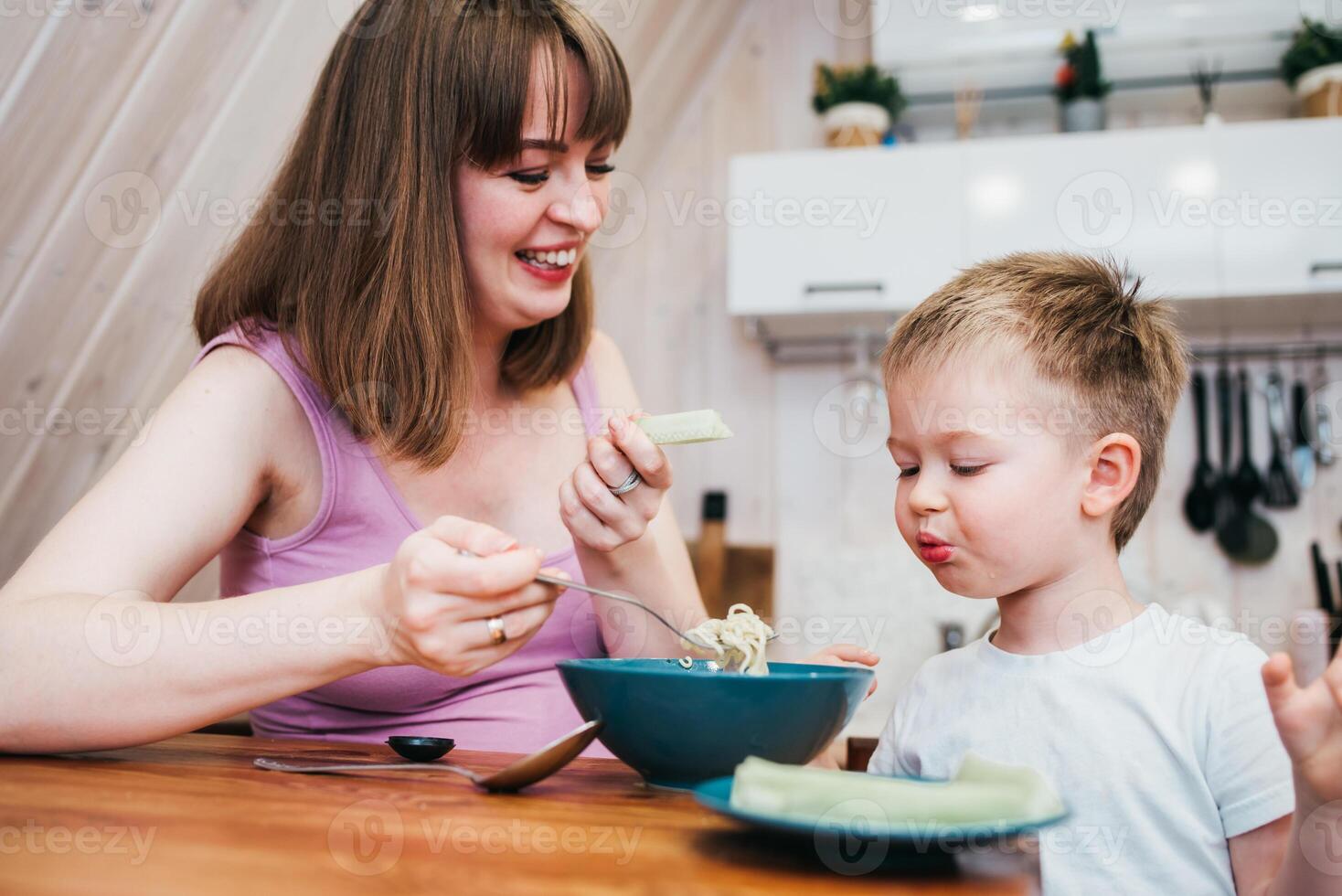 Cheerful child eating pasta in the kitchen with mom photo