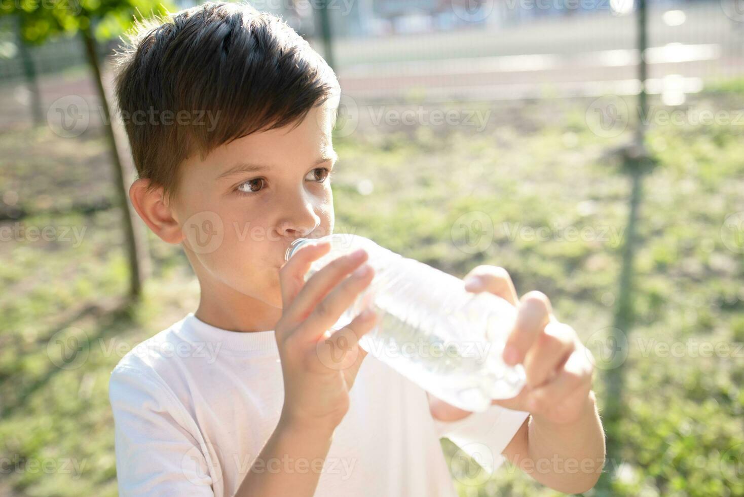 Cute boy sitting on the grass drinks water from a bottle in the summer at sunset. Child quenches thirst on a hot day photo