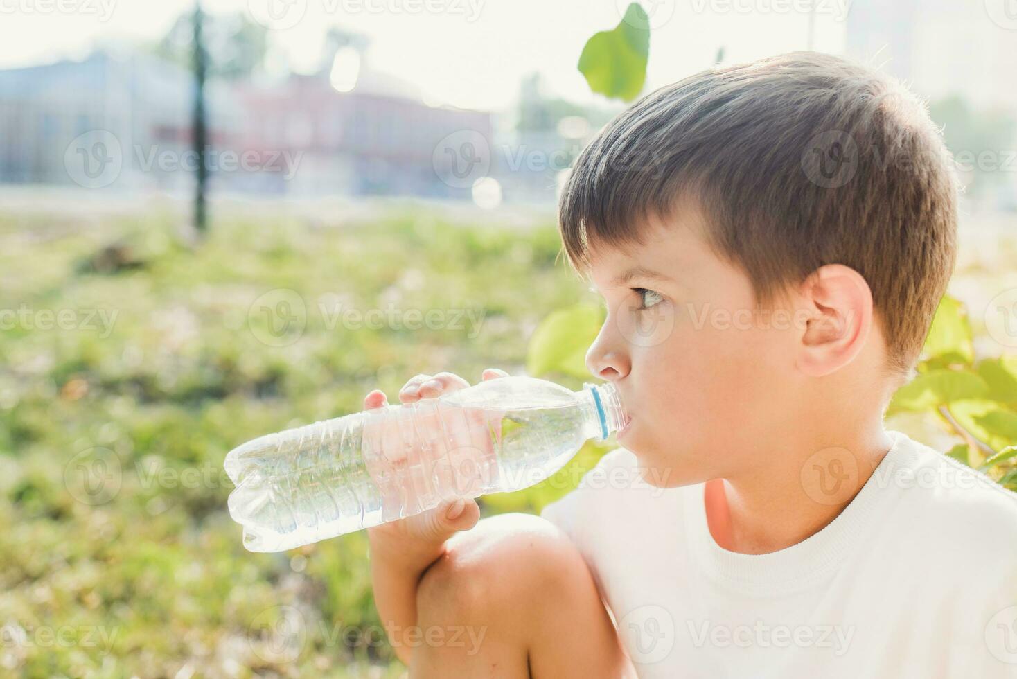 Cute boy sitting on the grass drinks water from a bottle in the summer at sunset. Child quenches thirst on a hot day photo