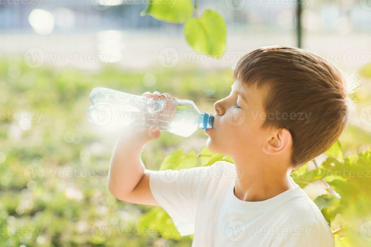 Cute boy sitting on the grass drinks water from a bottle in the summer at sunset. Child quenches thirst on a hot day photo