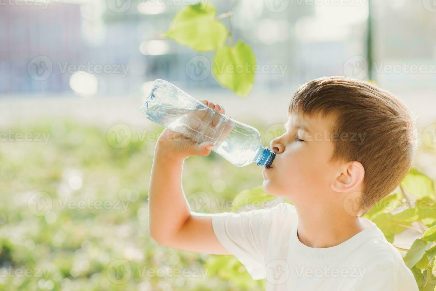 A beautiful child sitting on the grass drinks water from a bottle in the summer at sunset. Boy quenches his thirst on a hot day photo