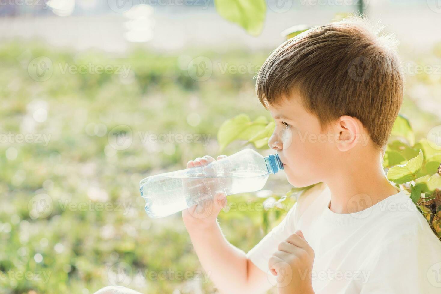 Cute boy sitting on the grass drinks water from a bottle in the summer at sunset. Child quenches thirst on a hot day photo