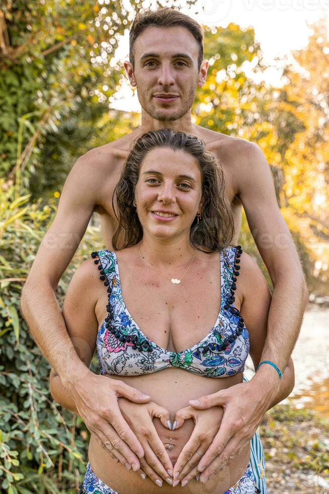 portrait of parents in swimsuits forming heart sign with hands on pregnant mom belly photo