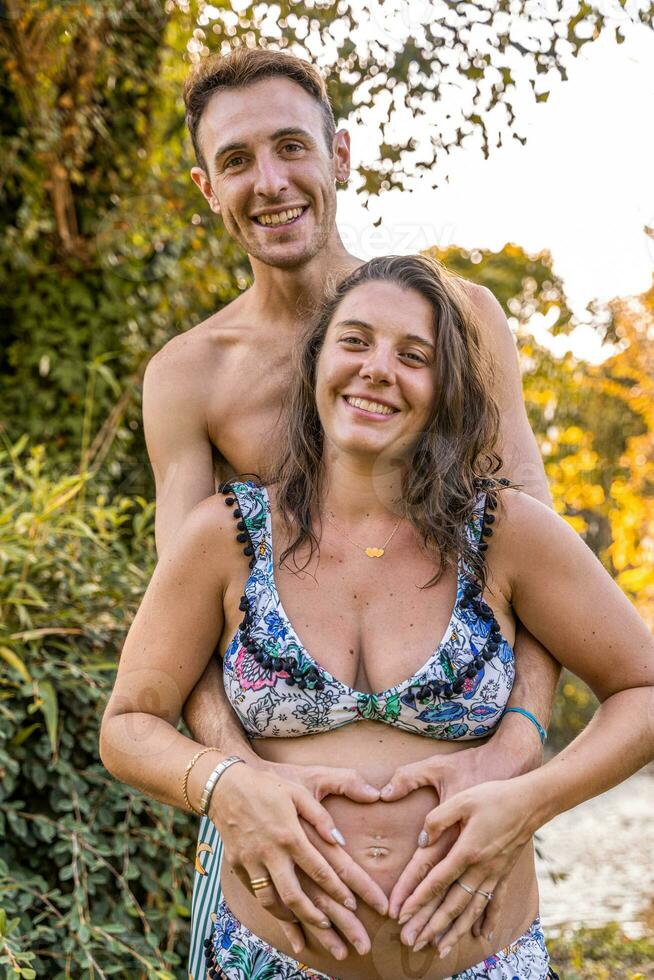 portrait of parents in swimsuits forming heart sign with hands on pregnant mom belly photo