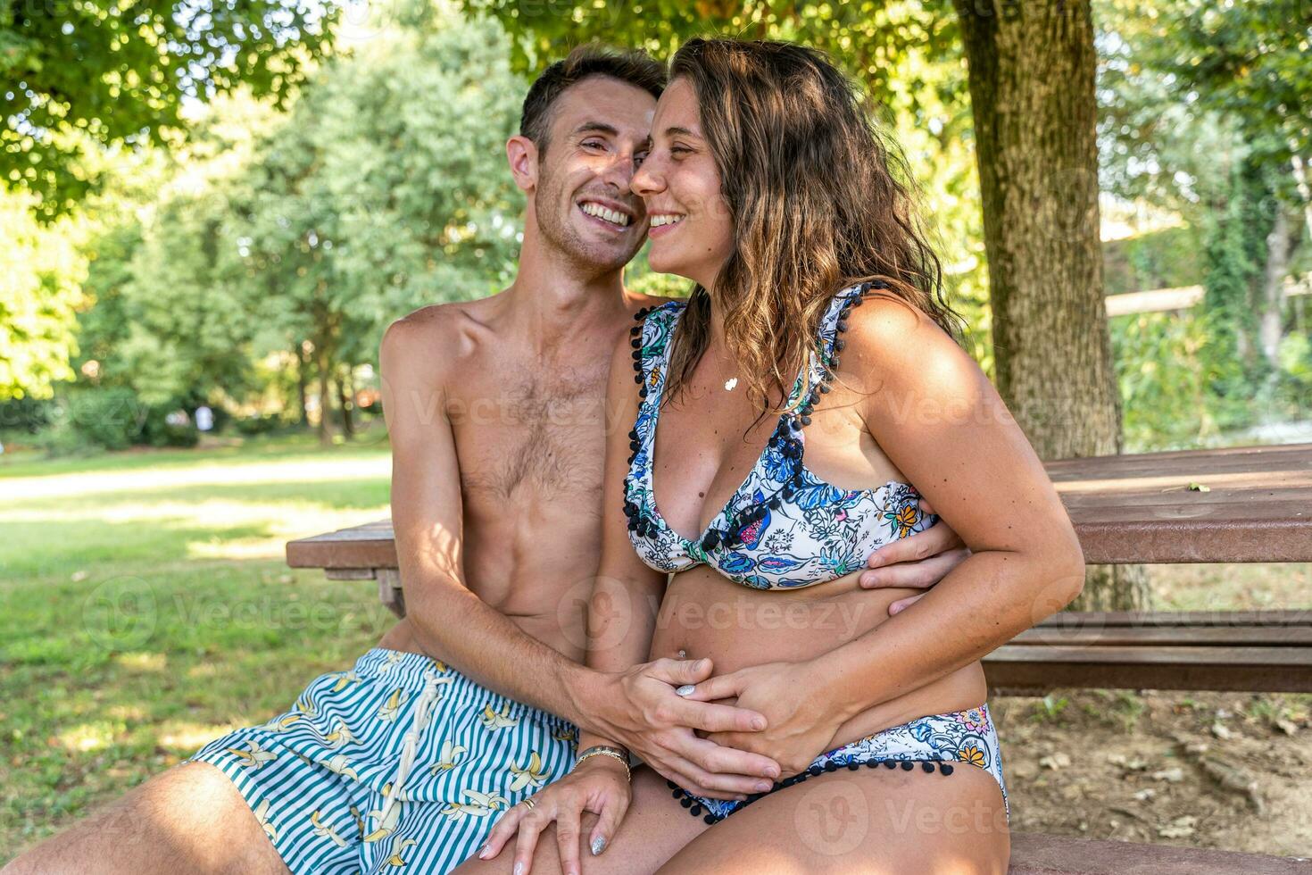 portrait of happy pregnant couple in swimwear kissing while sitting on a bench in a park photo