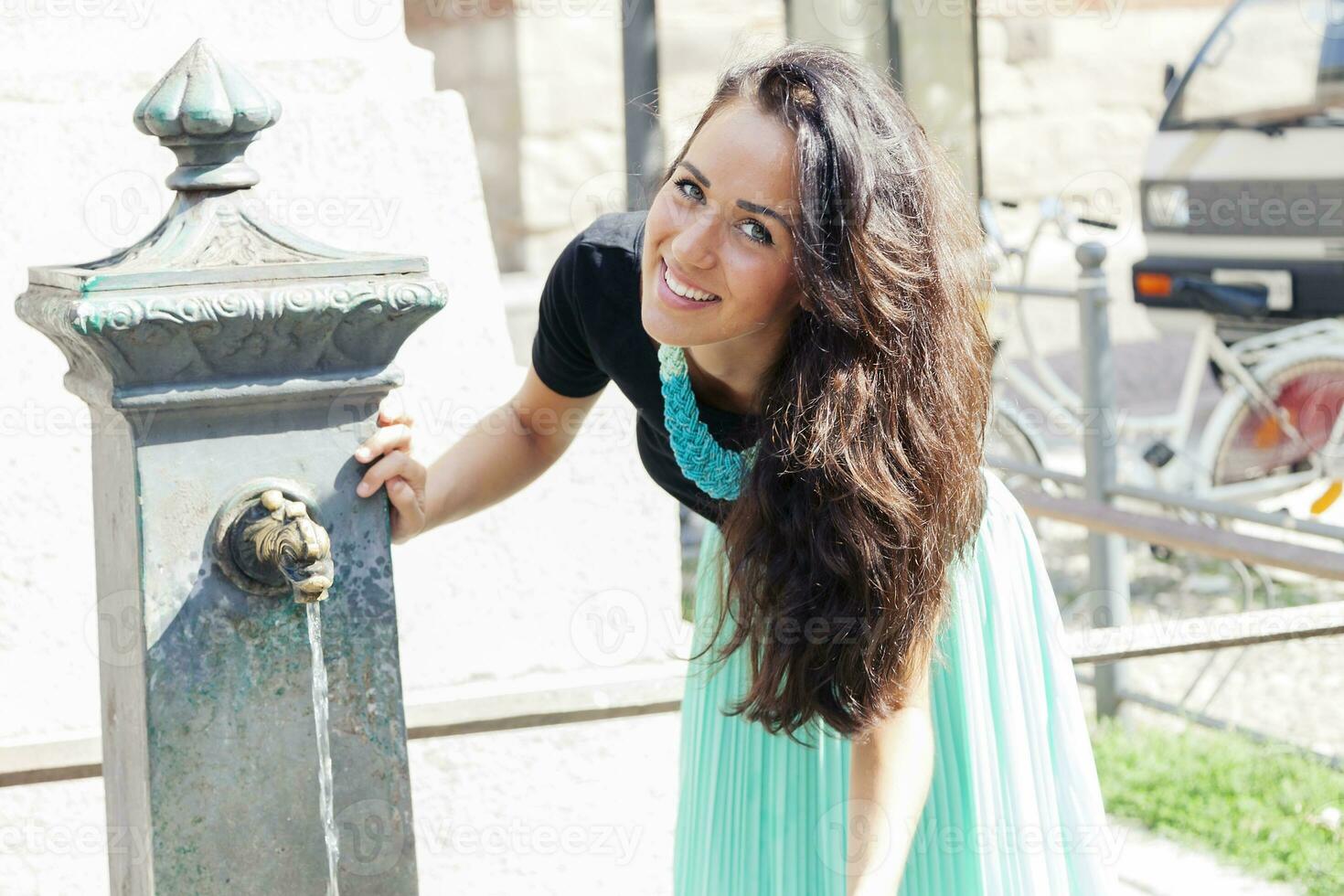 portrait of pretty girl drinks water from source in summer city photo