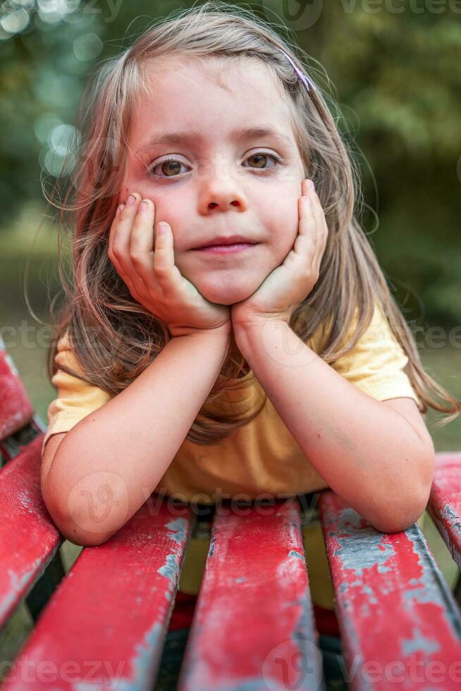 retrato de pequeño linda niña propensión con codos en un rojo banco al aire libre foto