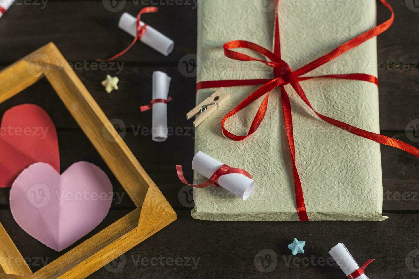 Flat lay romantic background. Giftbox, rolled wish papers, and wooden frame with paper hearts on dark wood board. photo