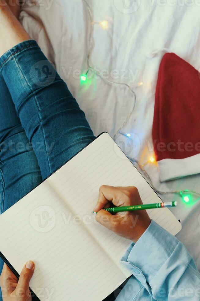 High angle view to young woman lying in bed and writing with pen on notebook. Wintertime. Christmas holidays. A woman making plans for the new year photo