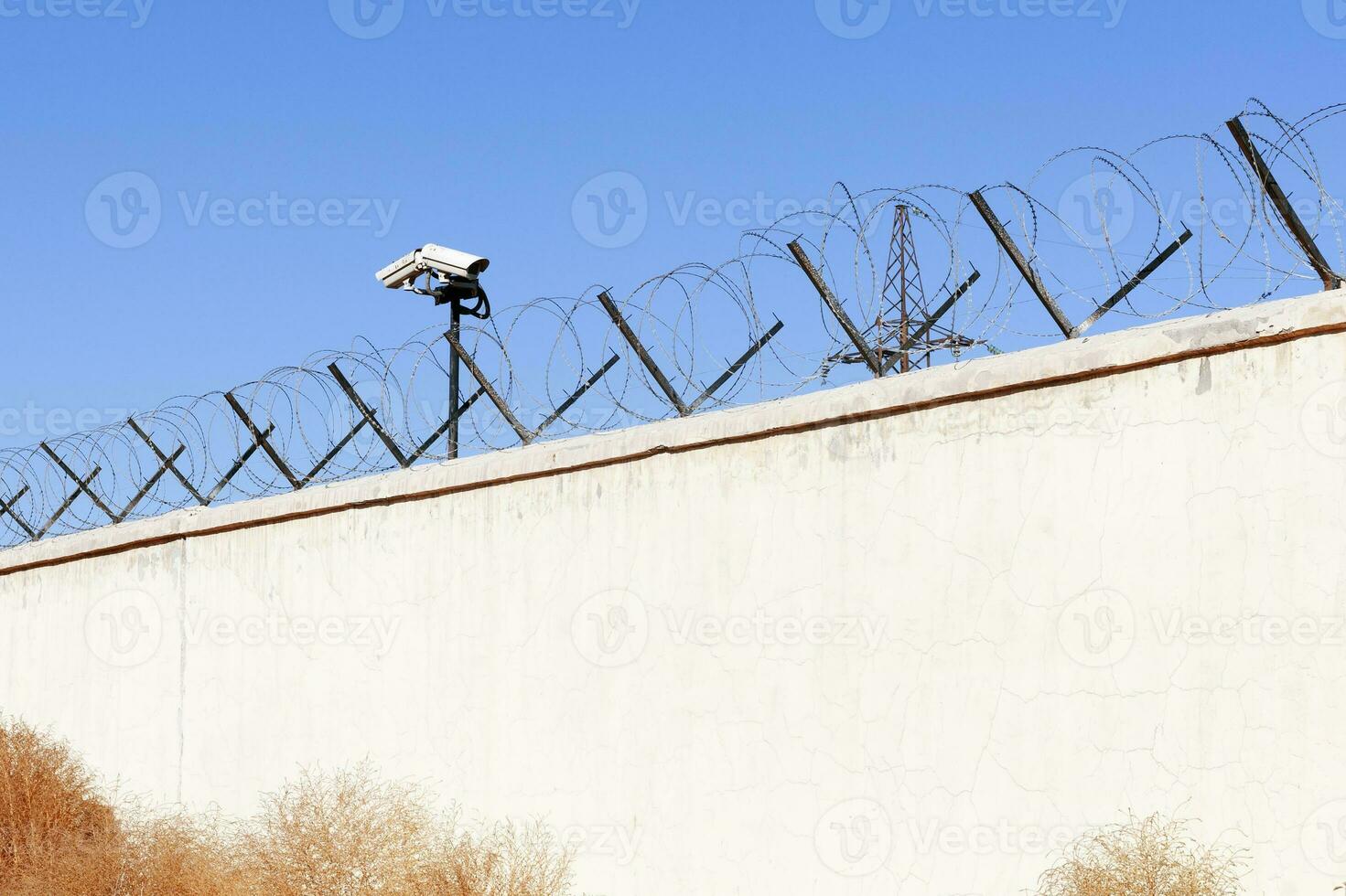 Security camera behind barbed wire fence stretched around prison walls. Security, surveillance camera watching area photo