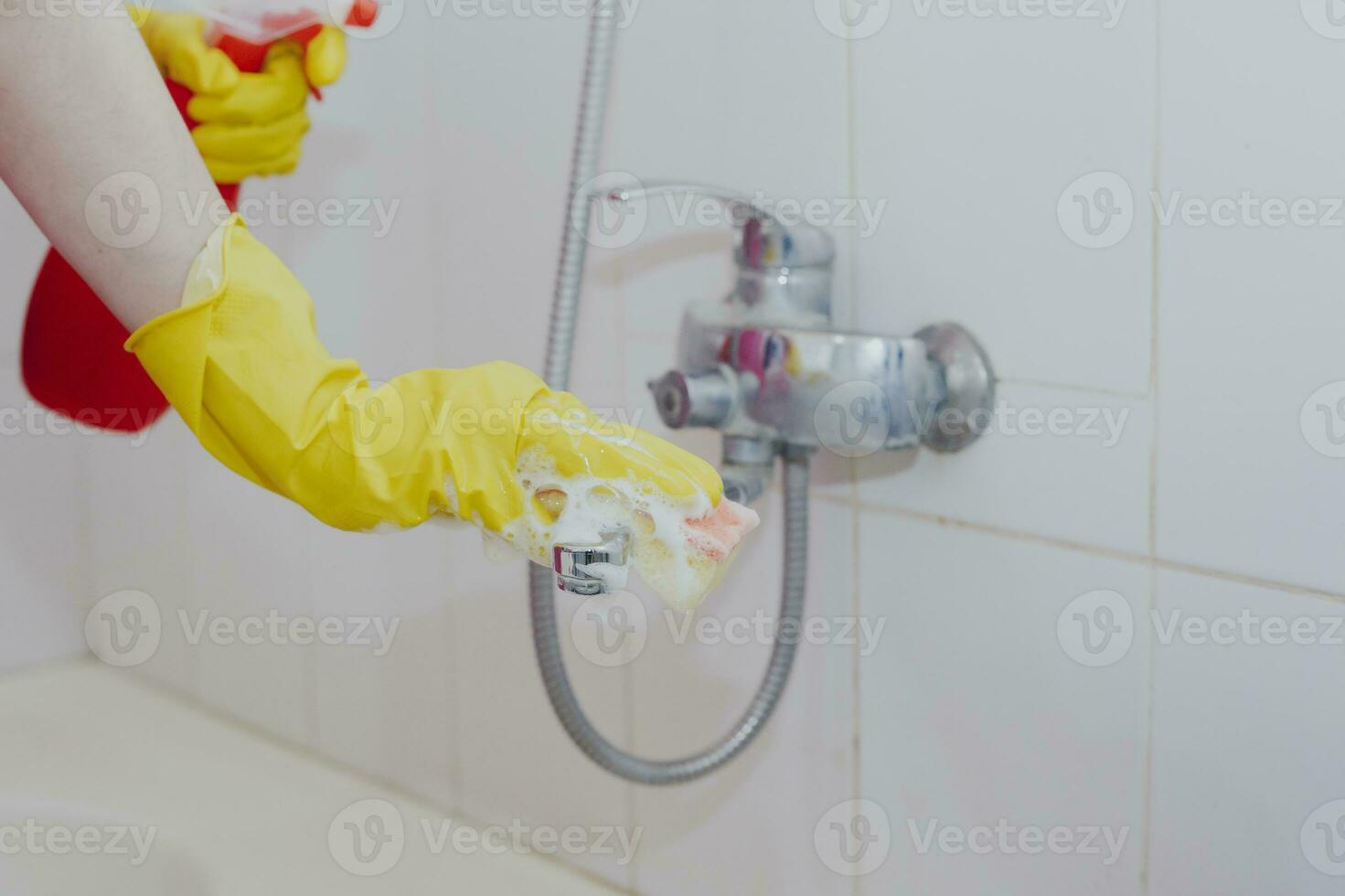 Housewife cleaning bathroom tap and shower Tap. Maid in yellow protective gloves washing dirty bath tap. Hands of woman washing bath photo