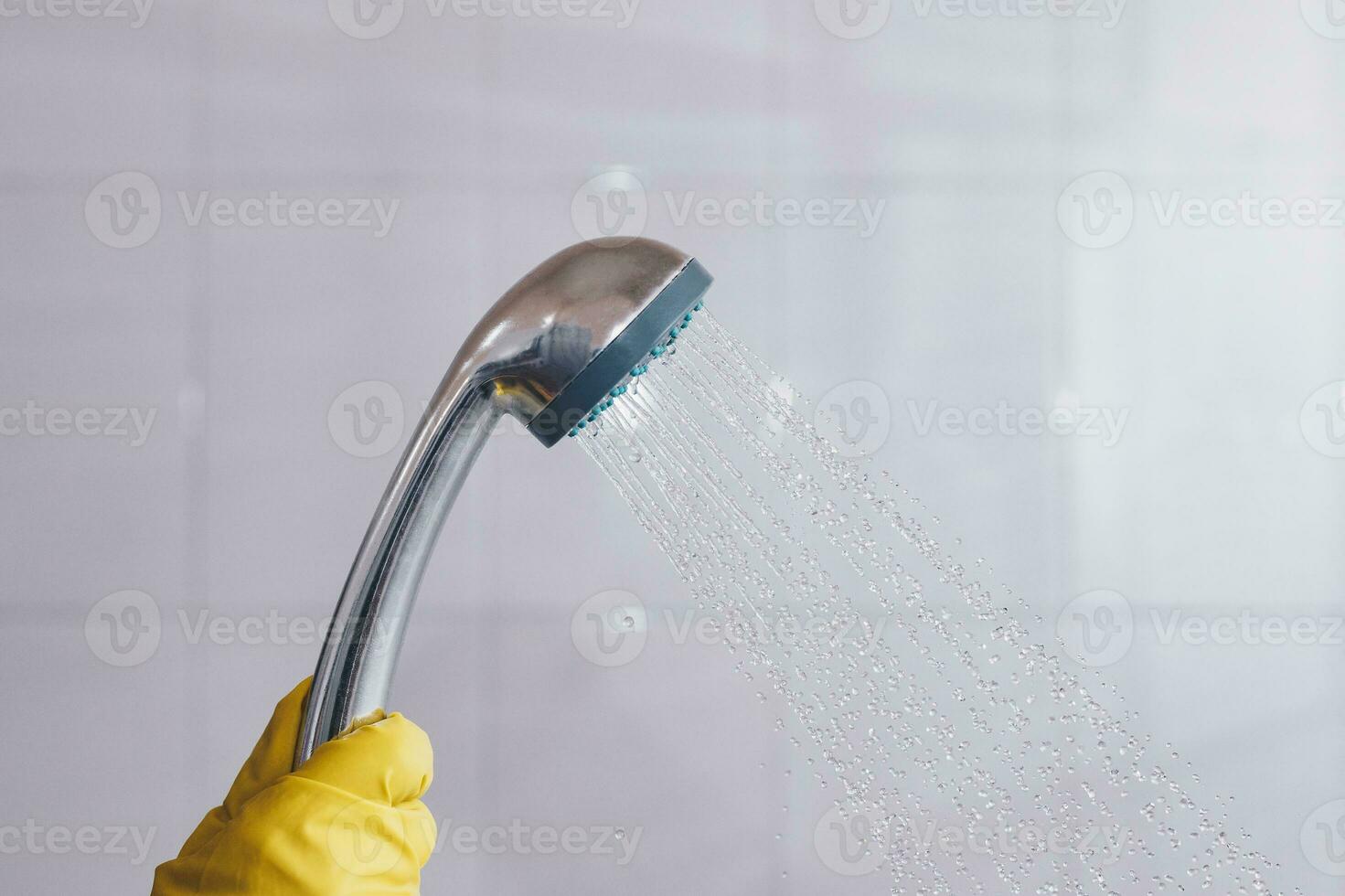 A person in rubber gloves holding showerhead with flowing water. Women in yellow protective gloves holding the head of the shower with running water in the bath photo