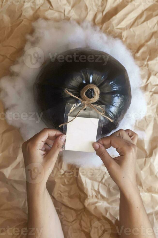 Woman preparing a halloween present. Hands prepare black pumpkin with empty white label tag photo