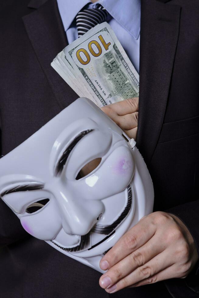 A mystrious person in formal wear holds anonymous mask and pile of us dollars photo