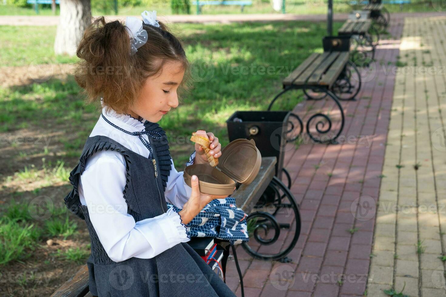 Girl with backpack eating sandwich packed in a sandwich box near school. A quick snack with a bun, unhealthy food, lunch from school. Back to school. Education, primary school classes, September 1 photo