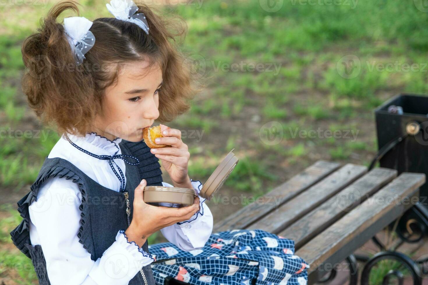 Girl with backpack eating sandwich packed in a sandwich box near school. A quick snack with a bun, unhealthy food, lunch from school. Back to school. Education, primary school classes, September 1 photo