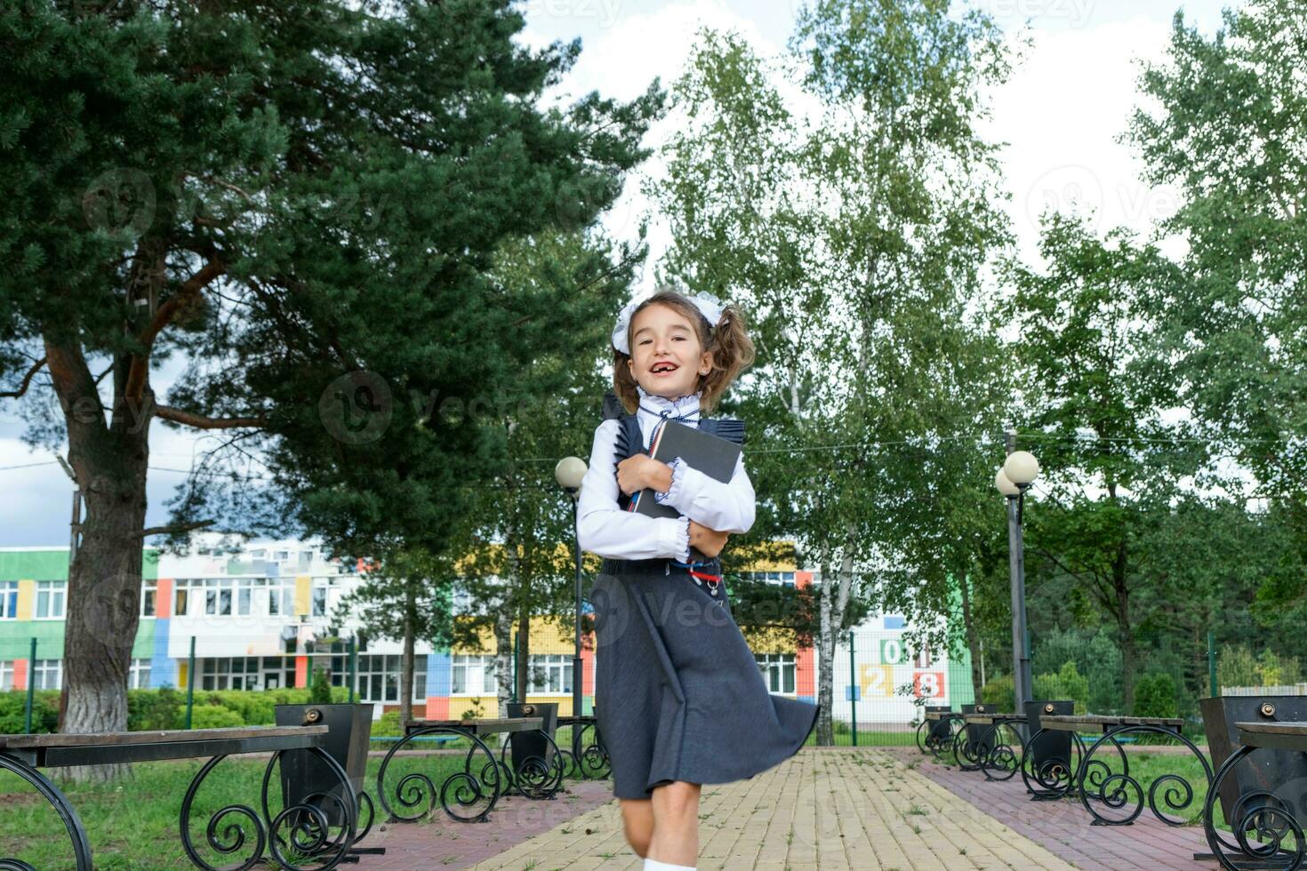 Cheerful funny girl with toothless smile in school uniform with white bows running in school yard. Back to school, September 1. Happy pupil with backpack. Primary education, elementary class. motion photo