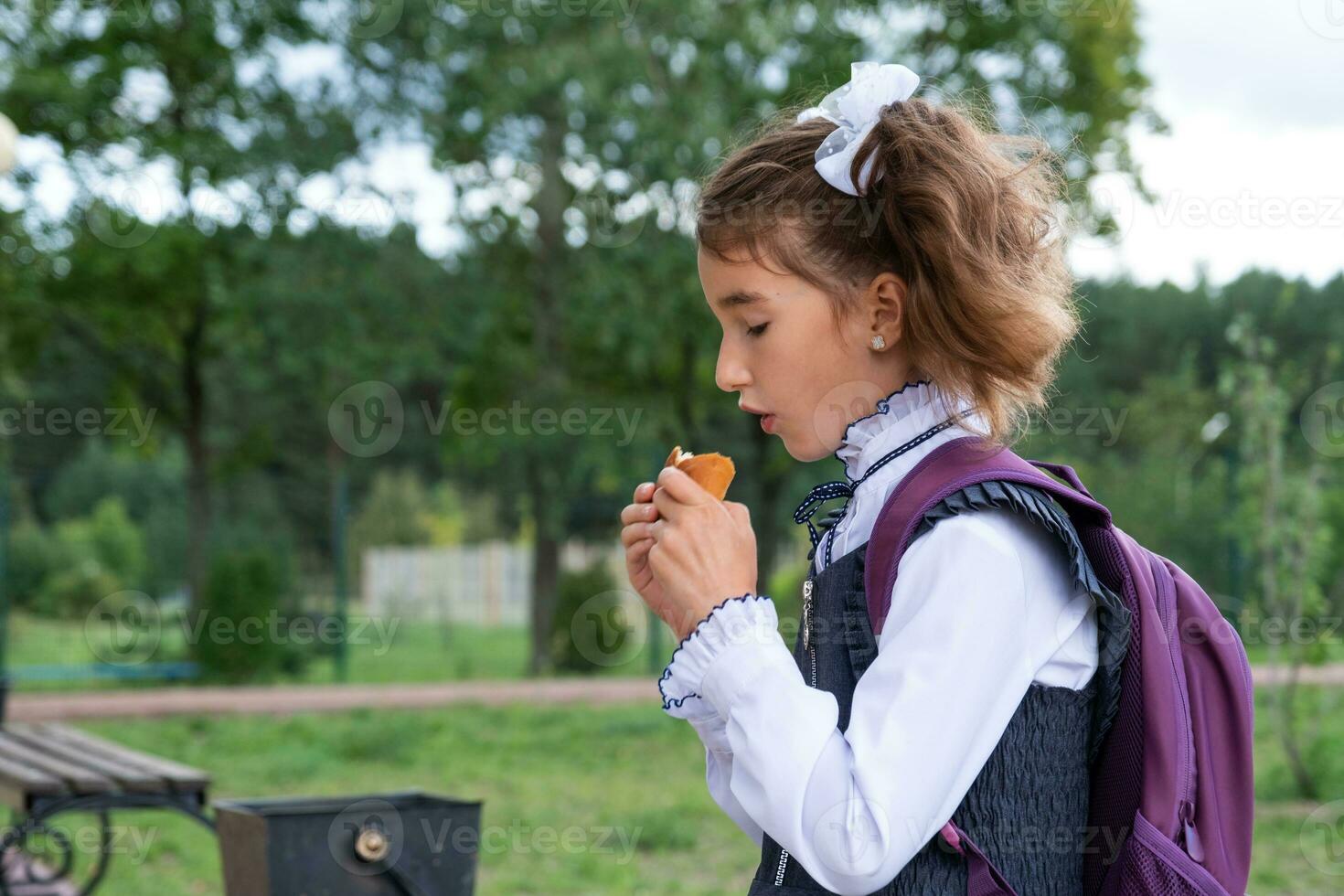 Girl with a backpack eating pie near school. A quick snack with a bun, unhealthy food, lunch from school. Back to school. Education, primary school classes, September 1 photo