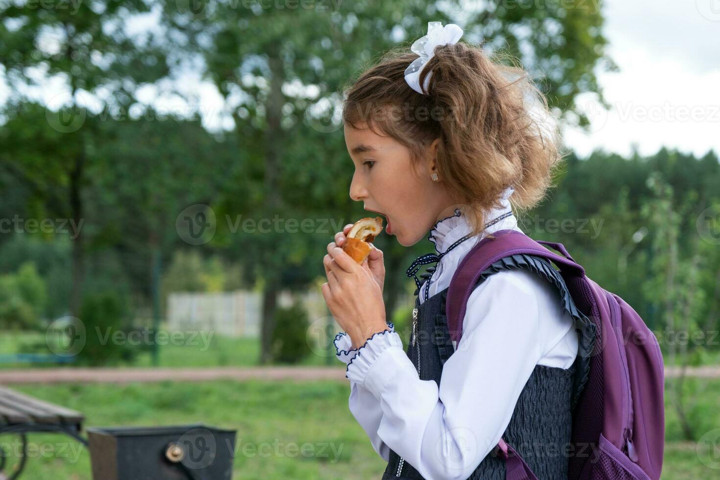 Girl with a backpack eating pie near school. A quick snack with a bun, unhealthy food, lunch from school. Back to school. Education, primary school classes, September 1 photo