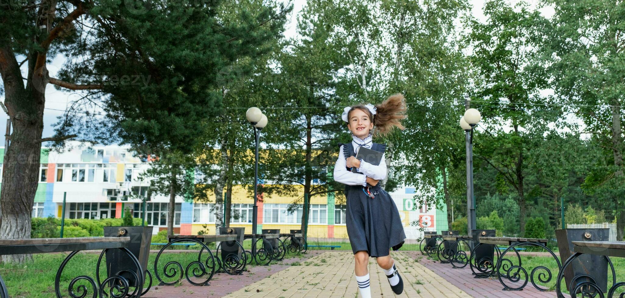 Cheerful funny girl with toothless smile in school uniform with white bows running in school yard. Back to school, September 1. Happy pupil with backpack. Primary education, elementary class. motion photo