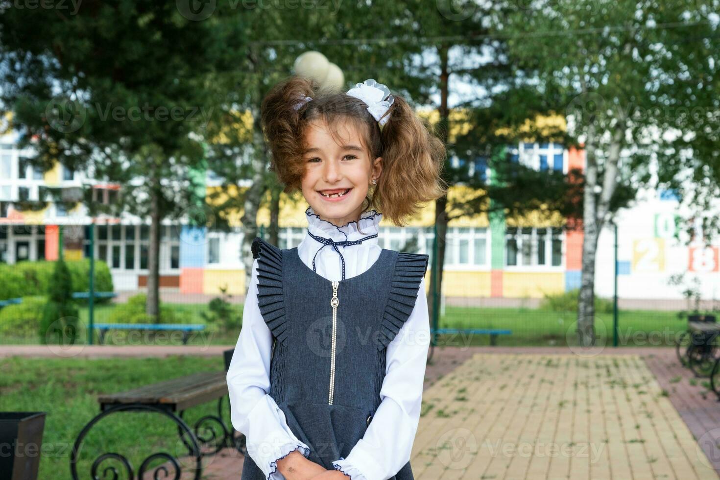 Cheerful funny girl with a toothless smile in a school uniform with white bows in school yard. Back to school, September 1. A happy pupil. Primary education, elementary class. Portrait of a student photo
