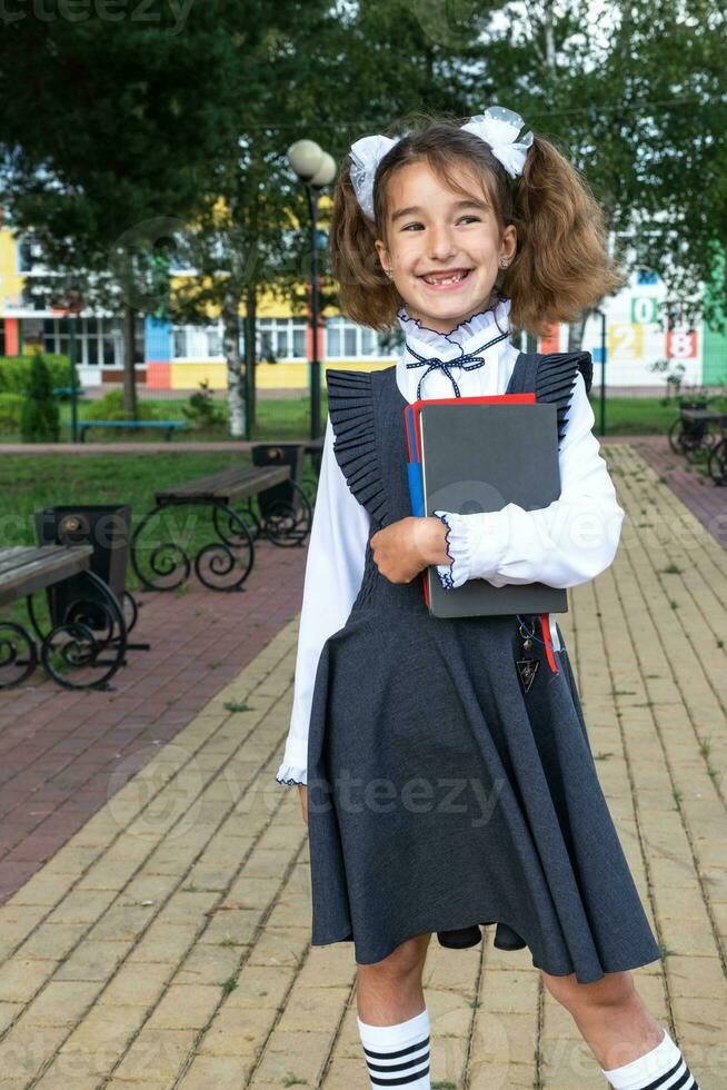 niña con mochila, colegio uniforme con blanco arcos y apilar de libros cerca escuela. espalda a escuela, contento alumno, pesado libros de texto educación, primario colegio clases, septiembre 1 foto