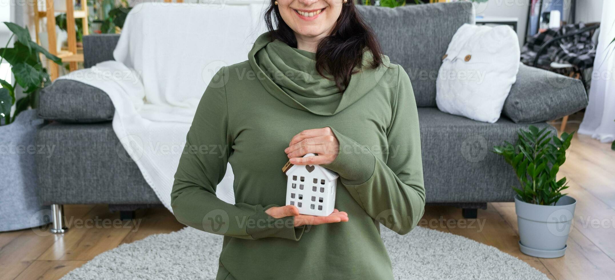 A happy woman in her house holds in her hands a miniature figure of a house and key in the interior. Dream house project, real estate purchase, insurance, mortgage, rent, reservation photo