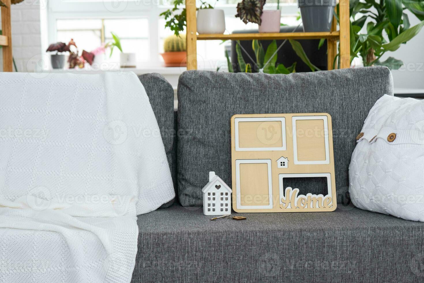 Key to home with keychain and mock up frame miniature of house on gray sofa in white interior with potted plants. Design, project, moving to new house, mortgage, rent and purchase real estate photo