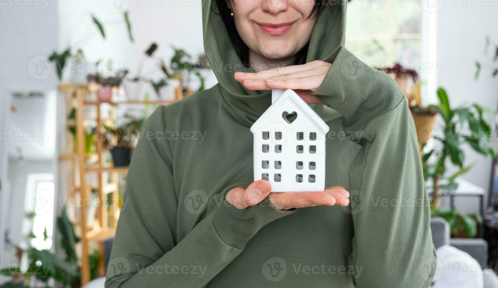 A happy woman in her house holds in her hands a miniature figure of a house and key in the interior. Dream house project, real estate purchase, insurance, mortgage, rent, reservation photo