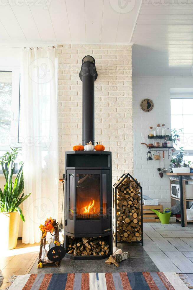 brillante soleado interior de el casa con negro metal acero hogar estufa con fuego y leña con Víspera de Todos los Santos decoración y otoño humor. acogedor hogar hogar en interior con interior en conserva planta foto