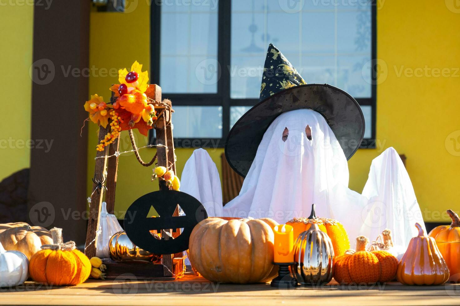 Child in bed sheet with slits like ghost in witch's hat and Halloween decor on the porch of the house outside in the yard of pumpkin, lantern, garlands, ack lantern. party, autumn mood photo