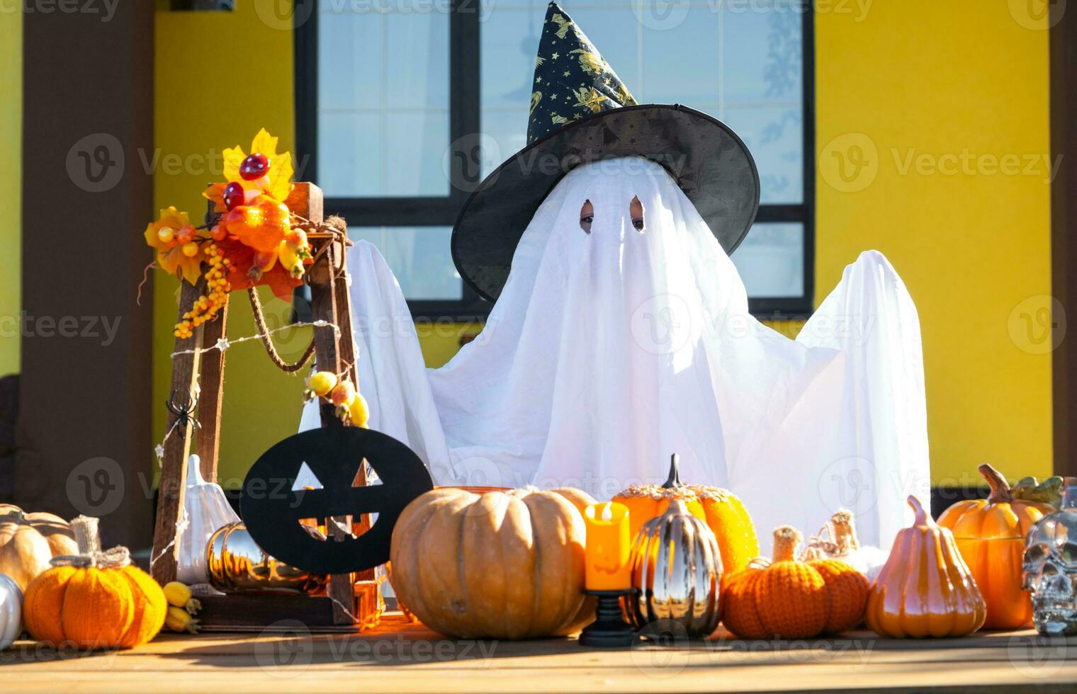 Child in bed sheet with slits like ghost in witch's hat and Halloween decor on the porch of the house outside in the yard of pumpkin, lantern, garlands, ack lantern. party, autumn mood photo