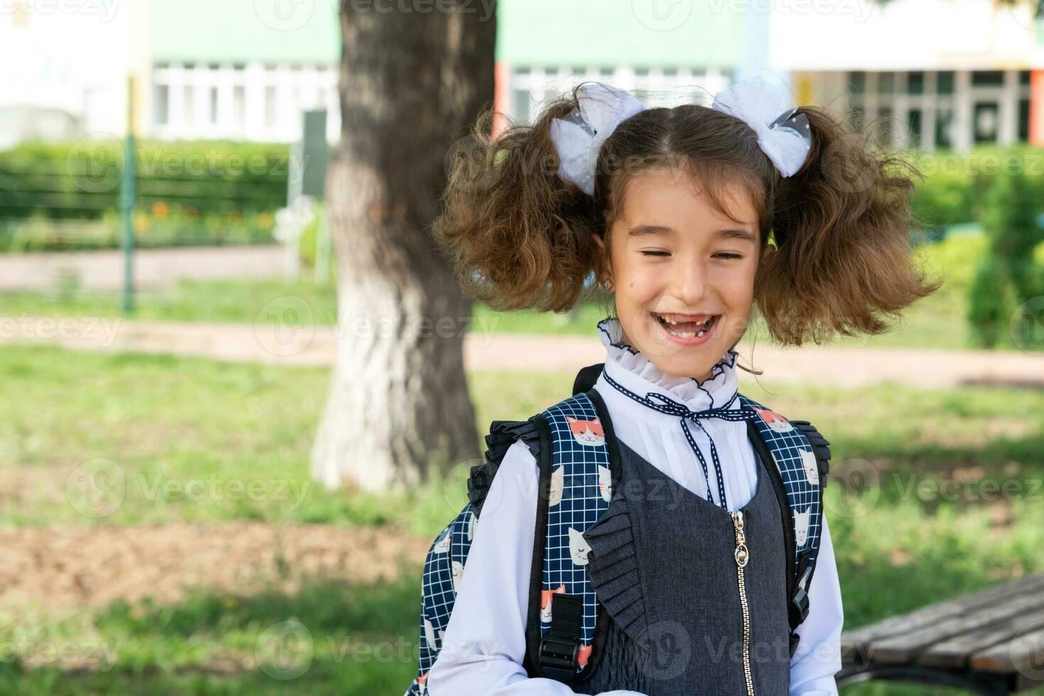 alegre gracioso niña con un sin dientes sonrisa en un colegio uniforme con blanco arcos en colegio patio trasero. espalda a escuela, septiembre 1. contento alumno con un mochila. primario educación, elemental clase. foto