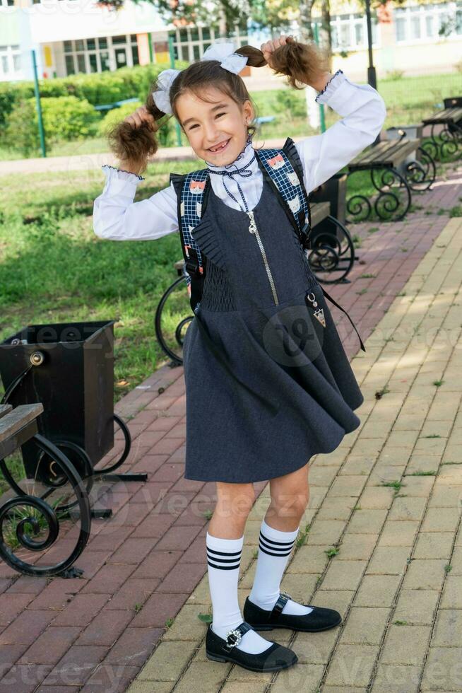 Cheerful funny girl with a toothless smile in a school uniform with white bows in school yard. Back to school, September 1. Happy pupil with a backpack. Primary education, elementary class. photo