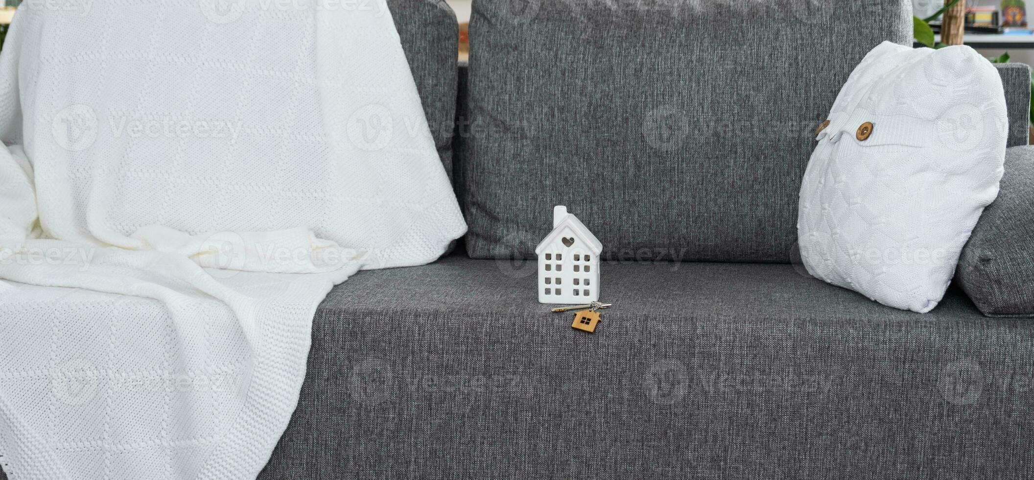 Key to home with keychain and and miniature of house on gray sofa in white interior with potted plants. Design, project, moving to new house, mortgage, rent and purchase real estate photo