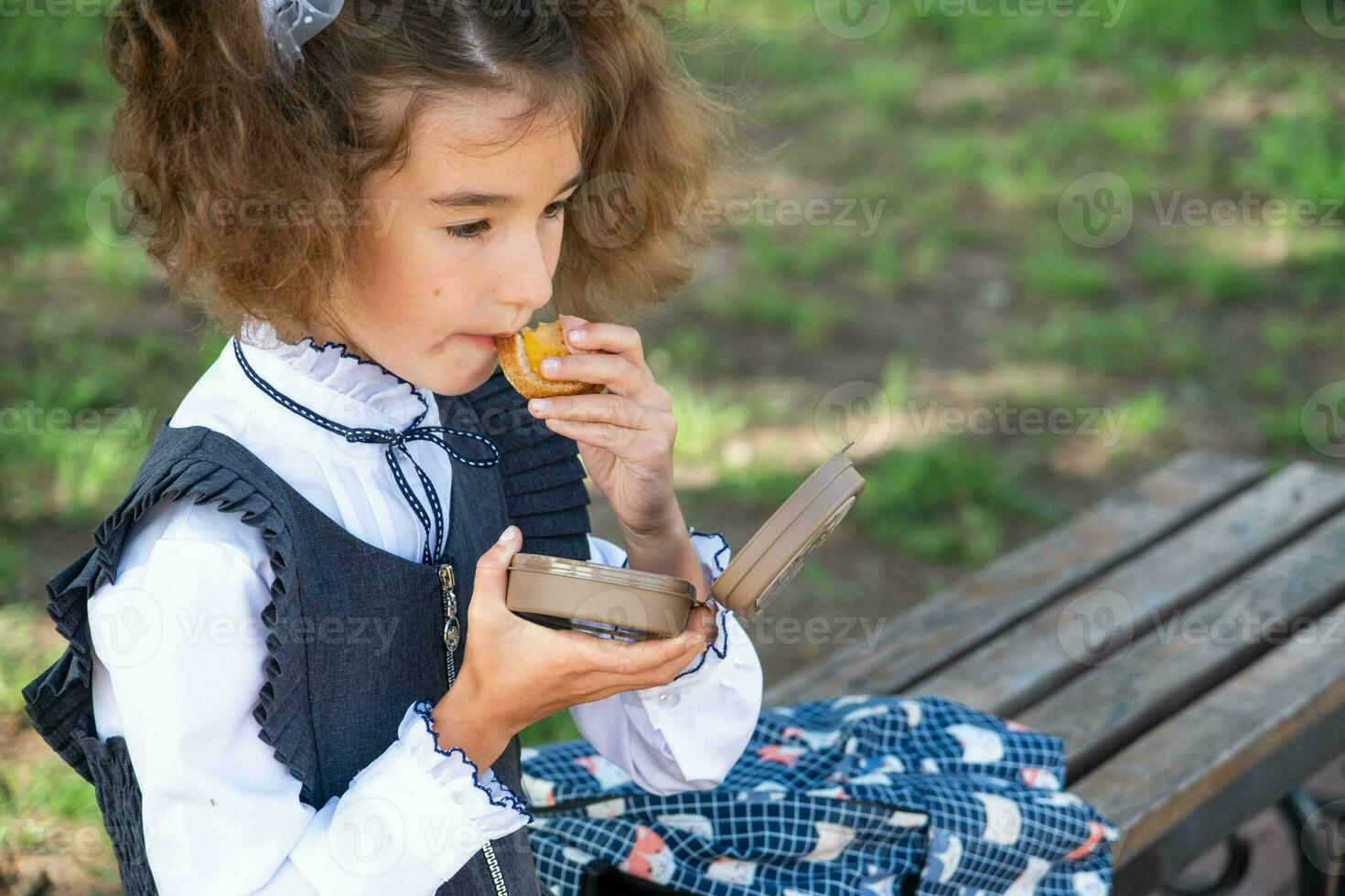 niña con mochila comiendo emparedado lleno en un emparedado caja cerca escuela. un rápido bocadillo con un bollo, insalubre alimento, almuerzo desde escuela. espalda a escuela. educación, primario colegio clases, septiembre 1 foto