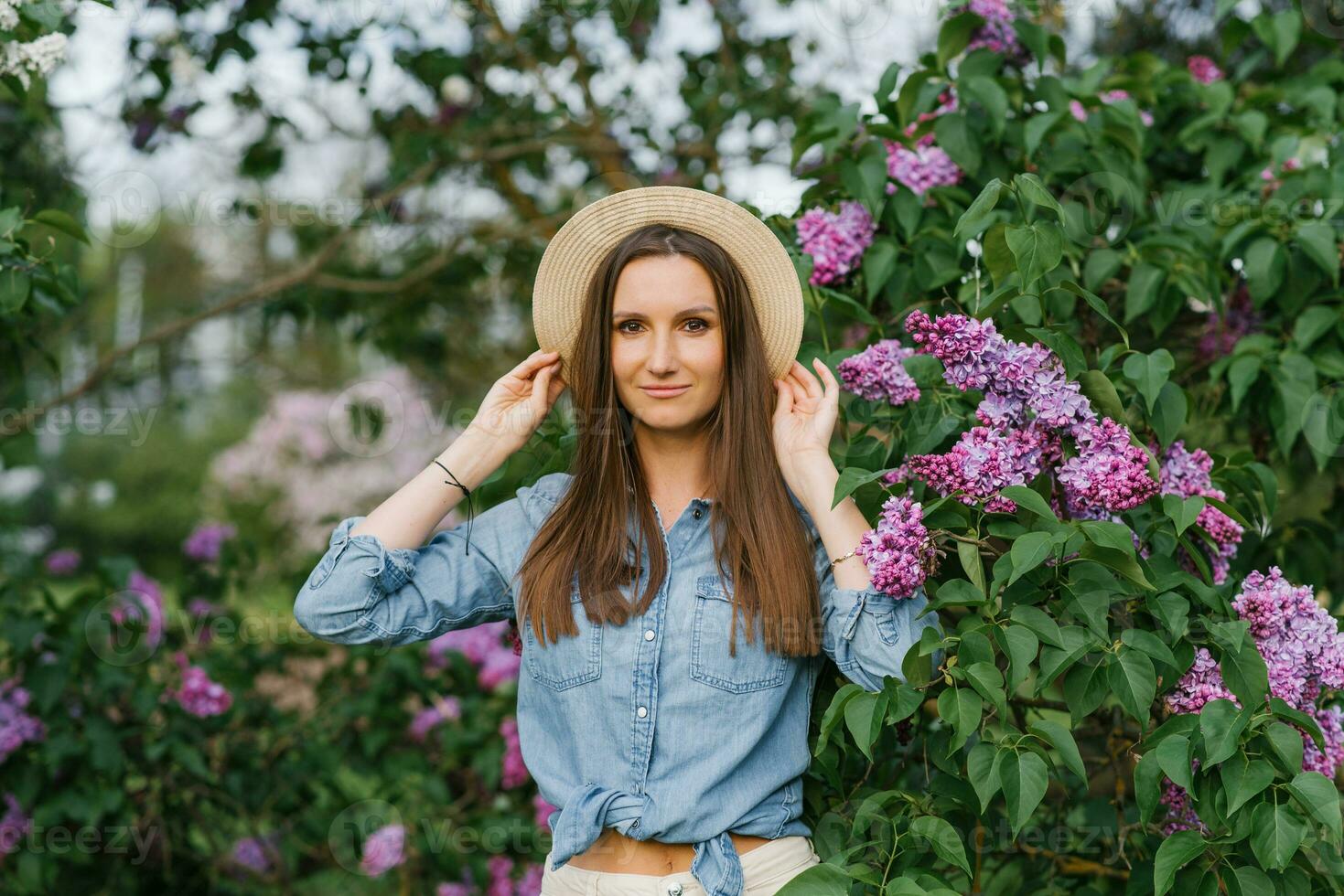 encantador joven mujer en un mezclilla camisa y un Paja sombrero camina entre el floreciente lilas en el primavera en el parque foto